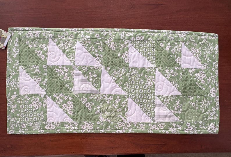 May include: A green and white quilted table runner with a floral pattern. The runner is made of fabric squares sewn together in a geometric pattern.