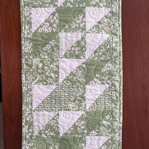 May include: A green and white table runner with a patchwork design. The runner is made of different fabrics with floral patterns and geometric designs. The runner is quilted with a white thread.