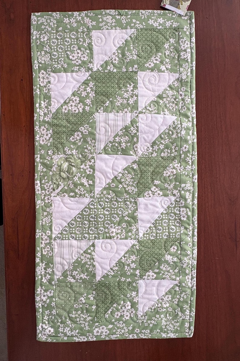 May include: A green and white table runner with a patchwork design. The runner is made of different fabrics with floral patterns and geometric designs. The runner is quilted with a white thread.