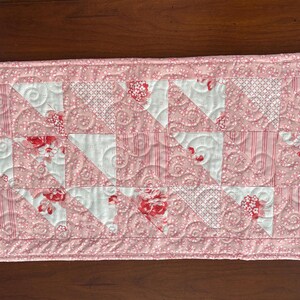 May include: A pink and white quilted table runner with a floral pattern. The runner is made of fabric squares sewn together in a geometric pattern.