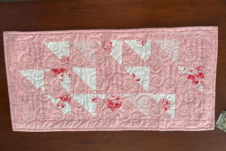 May include: A pink and white quilted table runner with a floral pattern. The runner is made of fabric squares sewn together in a geometric pattern.