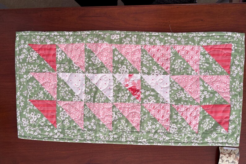 May include: A green and pink patchwork table runner with a floral pattern. The runner is made of fabric squares sewn together in a geometric pattern.