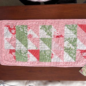 May include: A pink and green quilted table runner with a geometric pattern. The runner is made of fabric with floral prints and is approximately 16 inches wide and 48 inches long.