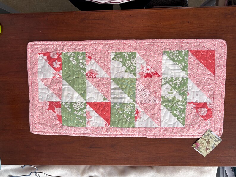 May include: A pink and green quilted table runner with a geometric pattern. The runner is made of fabric with floral prints and is approximately 16 inches wide and 48 inches long.