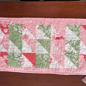 May include: A pink quilted table runner with a patchwork design featuring green, white, and red floral fabric squares. The runner has a pink border and is quilted with a small floral pattern.