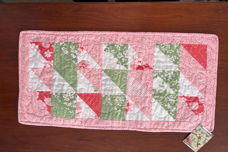 May include: A pink quilted table runner with a patchwork design featuring green, white, and red floral fabric squares. The runner has a pink border and is quilted with a small floral pattern.