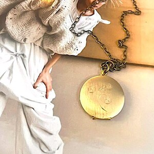 May include: A round, brushed gold-tone metal locket pendant featuring a subtle engraved floral design on the front surface. The pendant is suspended from a thick, textured, dark bronze-colored metal chain with large, interlocking oval links.