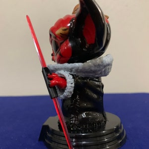 May include: A red and black Yoda figurine holding a red lightsaber. The figurine is wearing a black robe with a white fur collar. The figurine is standing on a black base.