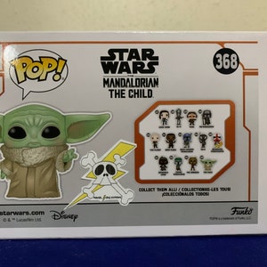May include: A Pop! Vinyl figure of The Child from the Star Wars: The Mandalorian series. The Child is a small, green alien with large ears and big eyes. The figure is wearing a brown robe and has a mischievous expression on its face. The figure is standing on a white background with a yellow and black skull and crossbones graphic behind it. The box also features a collection of other Pop! Vinyl figures from the Star Wars: The Mandalorian series.