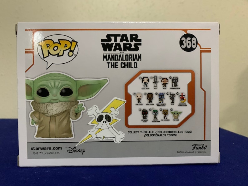 May include: A Pop! Vinyl figure of The Child from the Star Wars: The Mandalorian series. The Child is a small, green alien with large ears and big eyes. The figure is wearing a brown robe and has a mischievous expression on its face. The figure is standing on a white background with a yellow and black skull and crossbones graphic behind it. The box also features a collection of other Pop! Vinyl figures from the Star Wars: The Mandalorian series.