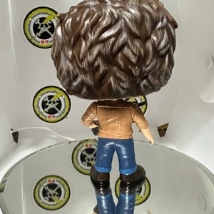 May include: A brown and white vinyl figure of a person wearing a brown jacket, blue jeans, and brown boots. The figure is holding a guitar in its right hand.