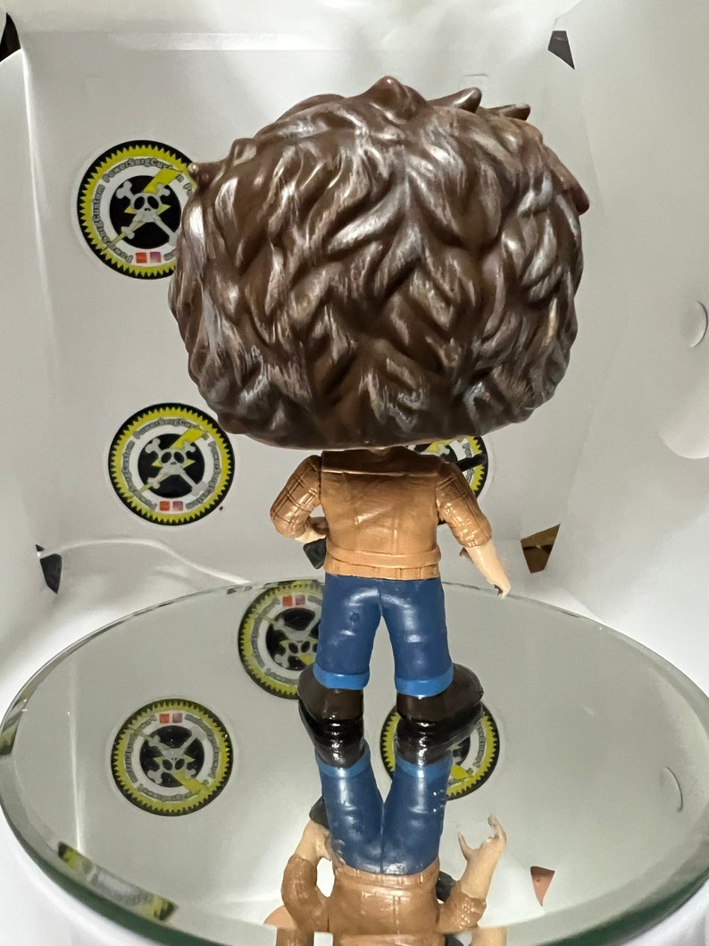 May include: A brown and white vinyl figure of a person wearing a brown jacket, blue jeans, and brown boots. The figure is holding a guitar in its right hand.