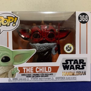 May include: A red and black Pop! Vinyl figure of The Child from Star Wars: The Mandalorian. The Child is wearing a red hooded robe and holding a small, silver object. The figure is in a clear plastic box with a white background. The box has the words "Pop!" and "The Child" on it, as well as the Star Wars logo and the Mandalorian logo.