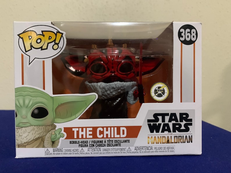 May include: A red and black Pop! Vinyl figure of The Child from Star Wars: The Mandalorian. The Child is wearing a red hooded robe and holding a small, silver object. The figure is in a clear plastic box with a white background. The box has the words "Pop!" and "The Child" on it, as well as the Star Wars logo and the Mandalorian logo.