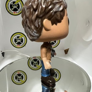 May include: A brown-haired pop culture figure wearing a brown and blue plaid shirt, blue jeans, and brown boots. The figure is standing on a white circular platform.