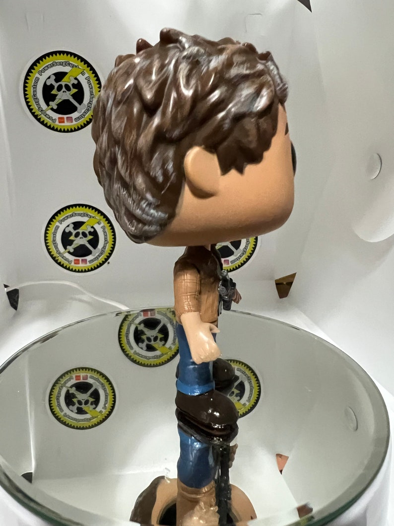 May include: A brown-haired pop culture figure wearing a brown and blue plaid shirt, blue jeans, and brown boots. The figure is standing on a white circular platform.