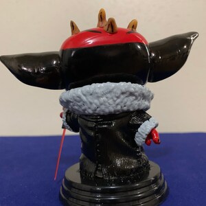 May include: A black and red painted figurine of a character with horns, wearing a black coat with a white fur collar and holding a red lightsaber. The figurine is standing on a black base.
