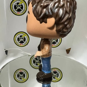 May include: A brown-haired figurine of a person wearing a brown jacket, blue jeans, and brown boots. The figurine is holding a small object in their right hand.