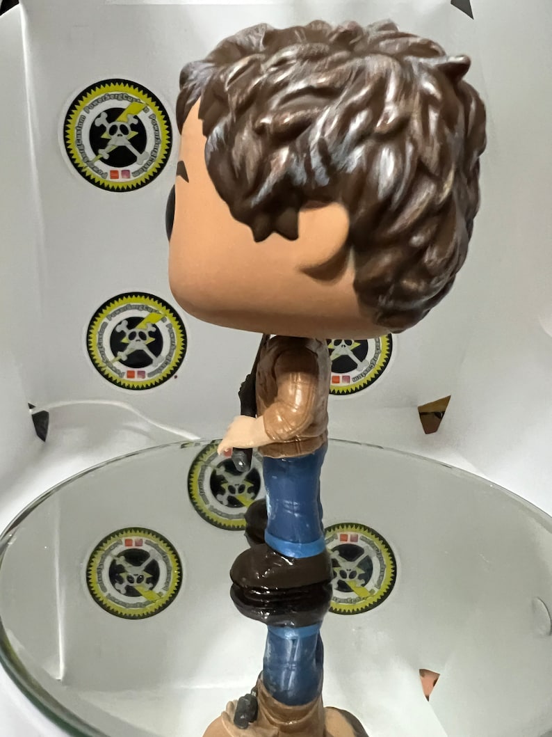 May include: A brown-haired figurine of a person wearing a brown jacket, blue jeans, and brown boots. The figurine is holding a small object in their right hand.