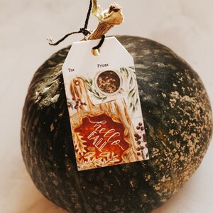 May include: A green pumpkin with a brown stem and a gift tag that says "Hello Fall" with a watercolor design of fall leaves and a cup of coffee. The tag also says "To" and "From".
