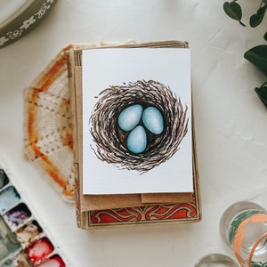 Op de afbeelding: Een aquarel wenskaart met een vogelnest met drie lichtblauwe eieren. De kaart ligt op een stapel kaarten in vintage-stijl met decoratieve randen. De afbeelding staat op een wit oppervlak met andere decoratieve items.