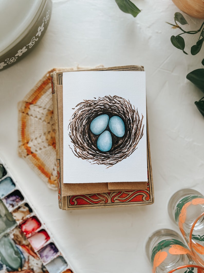 Op de afbeelding: Een aquarel wenskaart met een vogelnest met drie lichtblauwe eieren. De kaart ligt op een stapel kaarten in vintage-stijl met decoratieve randen. De afbeelding staat op een wit oppervlak met andere decoratieve items.