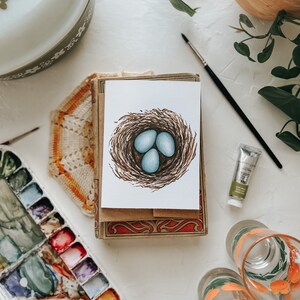 Op de afbeelding: Een aquarel van een vogelnest met drie blauwe eieren. Het kunstwerk staat op een witte kaart, rustend op een stapel kaarten met een decoratieve doos. Een palet met waterverf, een verftube en glazen met oranje ontwerpen zijn ook zichtbaar.