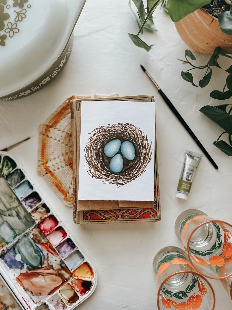 Op de afbeelding: Een aquarel van een vogelnest met drie blauwe eieren. Het kunstwerk staat op een witte kaart, rustend op een stapel kaarten met een decoratieve doos. Een palet met waterverf, een verftube en glazen met oranje ontwerpen zijn ook zichtbaar.