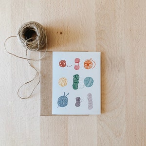 May include: A white card with colorful illustrations of yarn balls and knitting needles. The card is on a brown surface.
