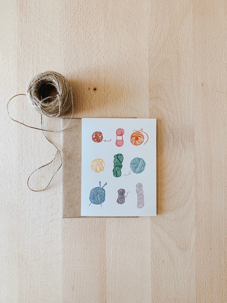 May include: A white card with colorful illustrations of yarn balls and knitting needles. The card is on a brown surface.