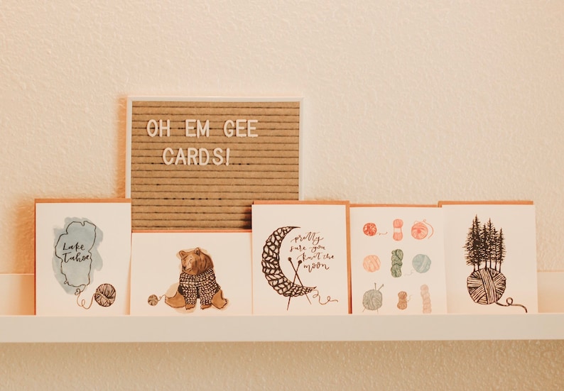 May include: A set of six greeting cards with illustrations of a bear wearing a sweater, a moon being knitted, a collection of yarn balls, and a group of trees growing out of a yarn ball. The cards are displayed on a white shelf with a sign that reads "Oh Em Gee Cards!"