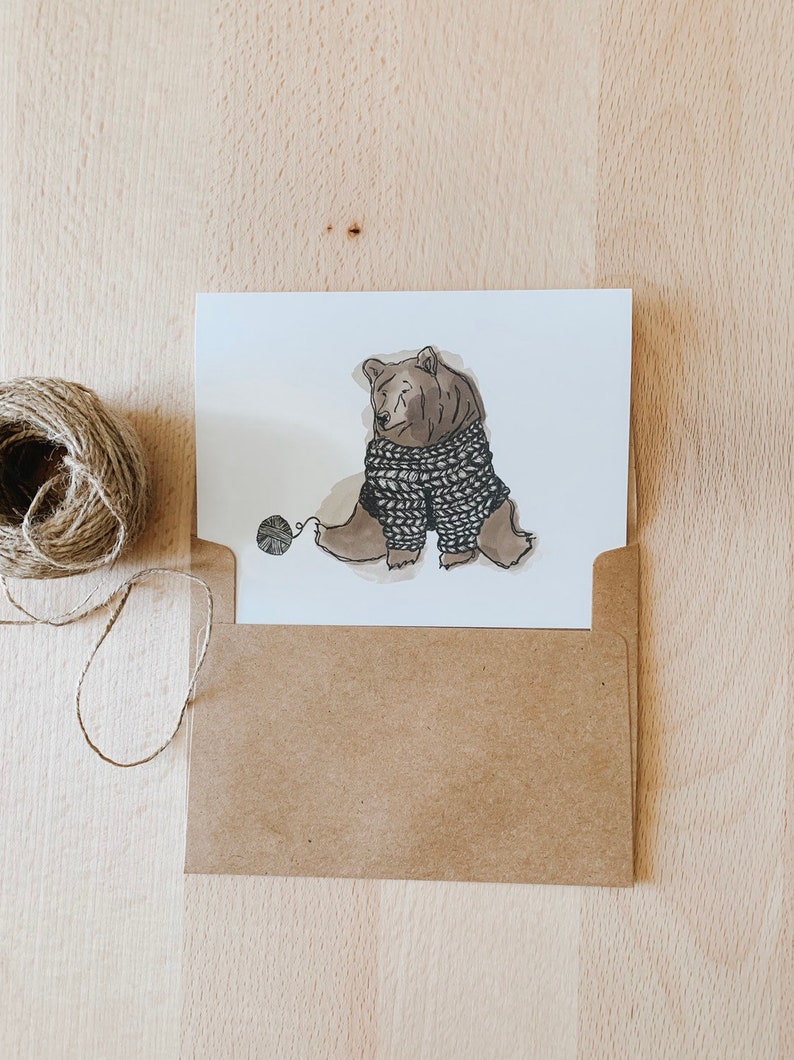 May include: A brown bear wearing a knitted sweater sits on a white background and holds a ball of yarn with its paw.