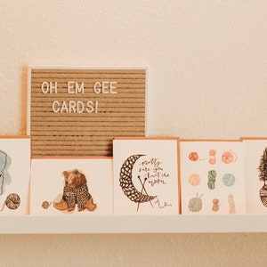 May include: A set of six greeting cards with illustrations of a bear wearing a sweater, a moon being knitted, a collection of yarn balls, and a group of trees growing out of a yarn ball. The cards are displayed on a white shelf with a sign that reads "Oh Em Gee Cards!"