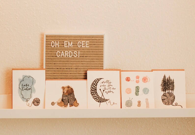 May include: A set of six greeting cards with illustrations of a bear wearing a sweater, a moon being knitted, a collection of yarn balls, and a group of trees growing out of a yarn ball. The cards are displayed on a white shelf with a sign that reads "Oh Em Gee Cards!"