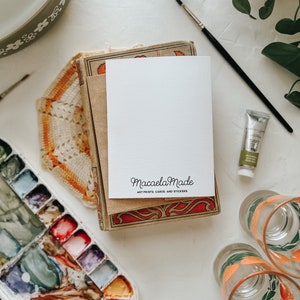 Op de afbeelding: Een stilleven met kunstbenodigdheden en briefpapier. Een witte kaart met de tekst "Macaela Made, Art Prints, Cards, and Stickers" ligt op een stapel vintage boeken. Een aquarelpalet, verftubes en glazen zijn ook zichtbaar.