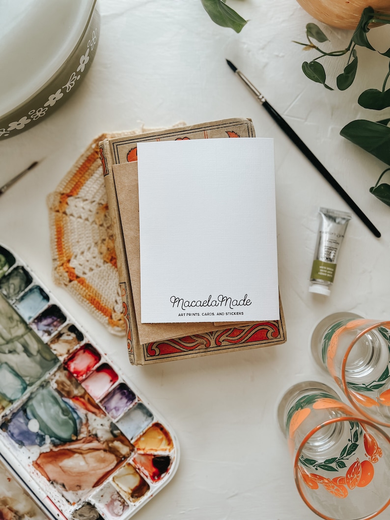 Op de afbeelding: Een stilleven met kunstbenodigdheden en briefpapier. Een witte kaart met de tekst "Macaela Made, Art Prints, Cards, and Stickers" ligt op een stapel vintage boeken. Een aquarelpalet, verftubes en glazen zijn ook zichtbaar.