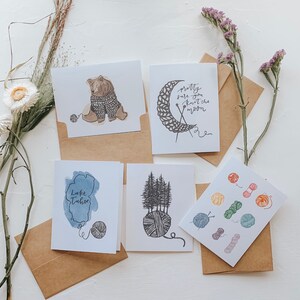 May include: Five greeting cards with different designs. The cards are all white with black ink. The designs include a bear wearing a sweater, a crescent moon with yarn, a lake, a group of trees, and a collection of yarn balls.
