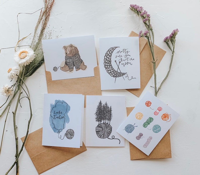 May include: Five greeting cards with different designs. The cards are all white with black ink. The designs include a bear wearing a sweater, a crescent moon with yarn, a lake, a group of trees, and a collection of yarn balls.