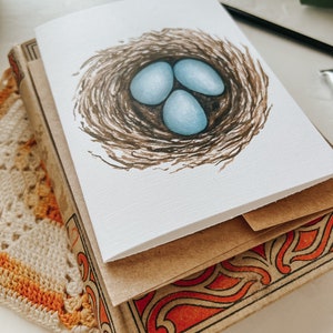 Op de afbeelding: Een wenskaart met een aquarel illustratie van drie lichtblauwe eieren in een bruin vogelnest. De kaart ligt op een stapel vintage boeken met decoratieve oranje en zwarte covers. Een gehaakt kleedje en een verfpalet zijn ook zichtbaar.