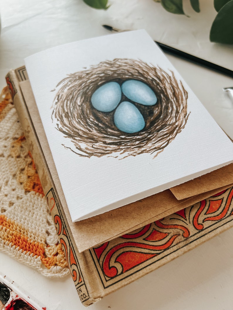 Op de afbeelding: Een wenskaart met een aquarel illustratie van drie lichtblauwe eieren in een bruin vogelnest. De kaart ligt op een stapel vintage boeken met decoratieve oranje en zwarte covers. Een gehaakt kleedje en een verfpalet zijn ook zichtbaar.