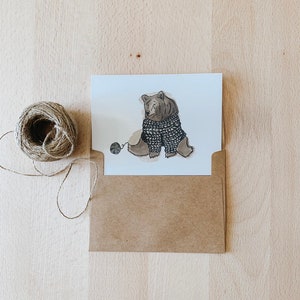 May include: A brown bear wearing a knitted sweater sits with a ball of yarn. The bear is drawn in a cartoon style with a simple background.