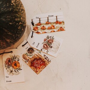 May include: Four gift tags with fall-themed designs. The tags are white with black text that says "To:" and "From:". The designs include pumpkins, a white pumpkin with flowers, and a wooden sign with the text "hello fall".