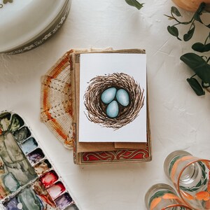 Op de afbeelding: Een wenskaart met een aquarel illustratie van een vogelnest en drie blauwe eieren. De kaart ligt op een stapel kaarten met een decoratieve rand. Andere items zijn een vintage ovenschaal, een aquarelpalet en decoratieve glazen.