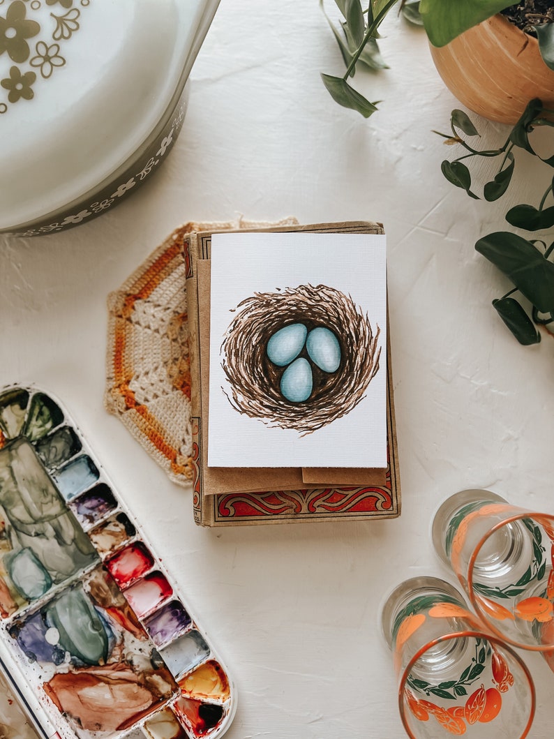 Op de afbeelding: Een wenskaart met een aquarel illustratie van een vogelnest en drie blauwe eieren. De kaart ligt op een stapel kaarten met een decoratieve rand. Andere items zijn een vintage ovenschaal, een aquarelpalet en decoratieve glazen.