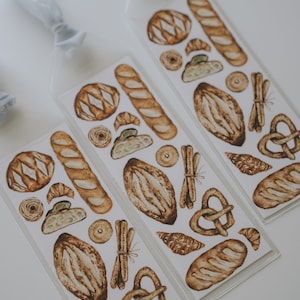 May include: Three clear plastic bookmarks with a white background and brown watercolor illustrations of various breads, pretzels, and pastries.