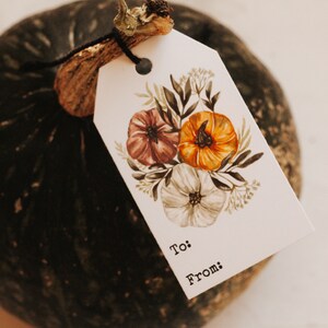 May include: A white gift tag with a floral design featuring pink, orange, and white flowers. The tag has the words "To:" and "From:" printed on it. The tag is tied to a green pumpkin.