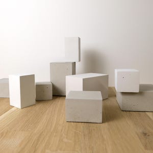 May include: A collection of geometric blocks in varying shades of white and light gray, arranged in a cluster. The set includes several rectangular prisms and cubes with smooth, solid surfaces, showcasing a mix of matte white and textured gray concrete.