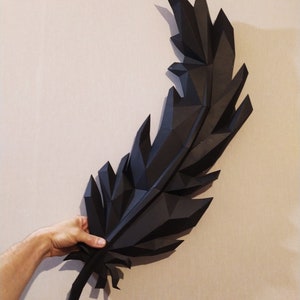 May include: A large black origami feather, folded from paper, is held in a person's hand. The feather is made up of many small triangles, giving it a geometric look.