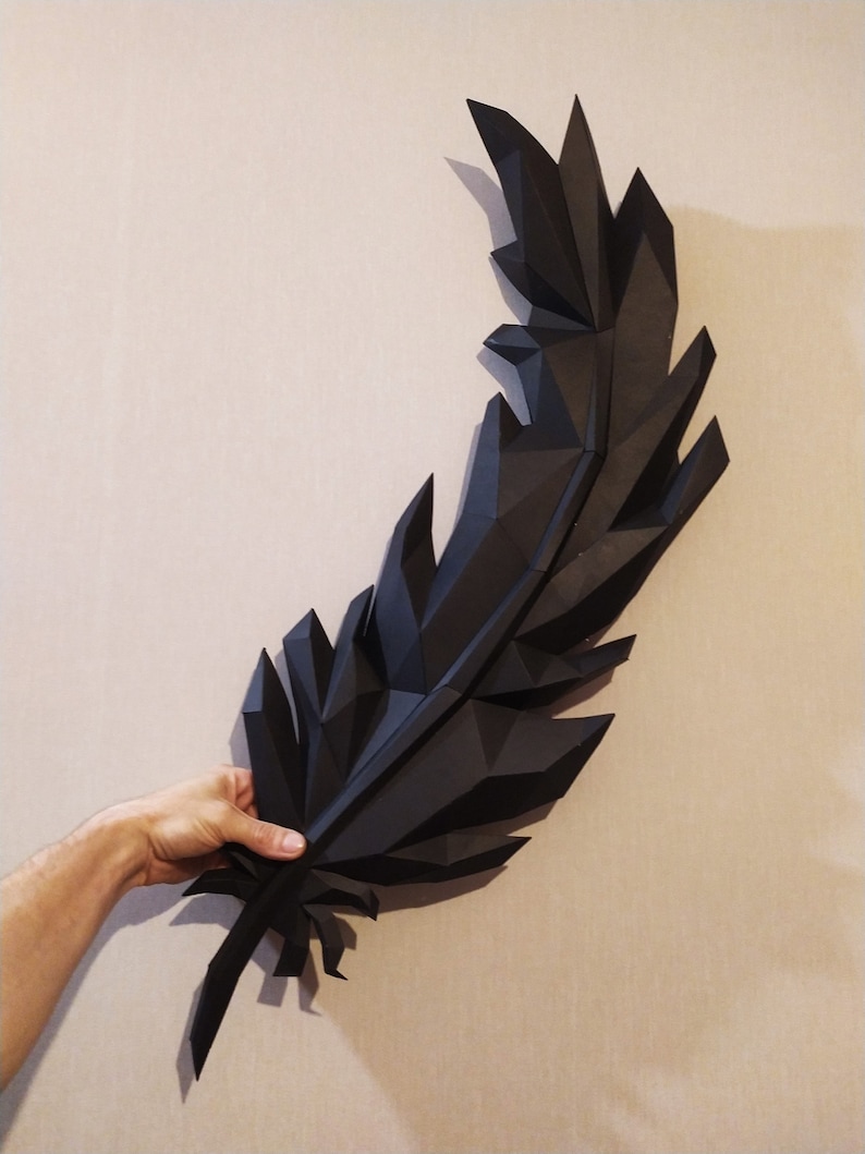 May include: A large black origami feather, folded from paper, is held in a person's hand. The feather is made up of many small triangles, giving it a geometric look.