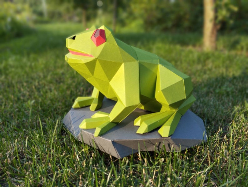 May include: A green origami frog with red eyes sitting on a gray rock. The frog is made of paper and has a geometric design.